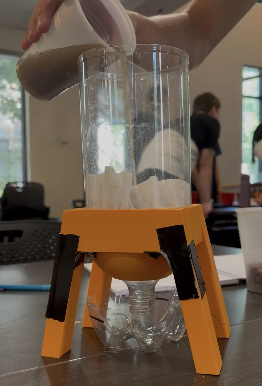 Muddy water being poured into a homemade filter: a clear plastic cylinder lined with filter paper, sitting on an orange 3D-printed stand over a cut plastic bottle